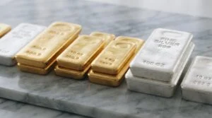 Close-up detalhado de barras físicas de ouro e prata posicionadas lado a lado sobre uma superfície de mármore cinza, com reflexos suaves que evidencia
